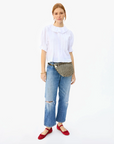 A woman stands against a white background, wearing a white blouse, distressed jeans, red flats, and a Clare Vivier Grande Fanny Woven Checker shoulder bag.