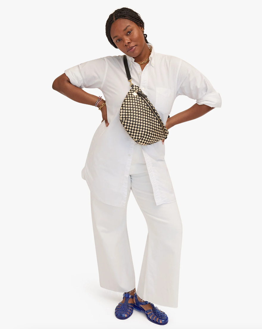 A confident woman stands with hands on hips, dressed in a white shirt and pants, accessorized with a Clare Vivier Grande Fanny Woven Checker bag and blue sandals, against a plain background. Her style exudes effortless elegance.