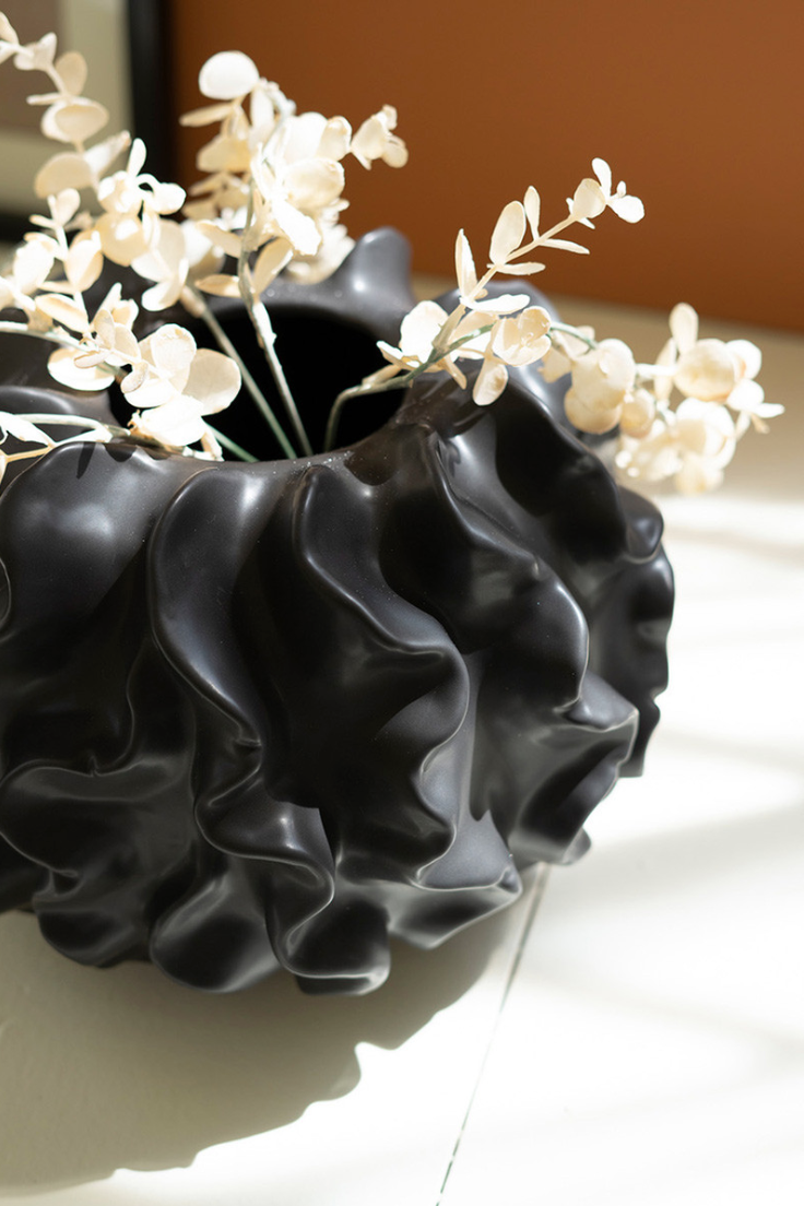 A Black Coral Ceramic Vase by Kalalou, Inc shaped like a human head with wavy hair, filled with delicate white flowers. The vase, embodying Arizona style, is illuminated by natural light, casting shadows on a beige background.