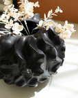 A Black Coral Ceramic Vase by Kalalou, Inc shaped like a human head with wavy hair, filled with delicate white flowers. The vase, embodying Arizona style, is illuminated by natural light, casting shadows on a beige background.