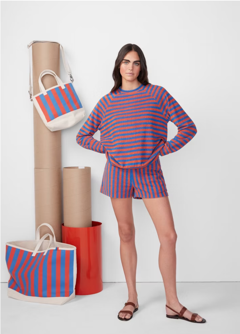A woman in a striped blue and red raglan sleeve Terry Franny sweatshirt and shorts stands beside two large tote bags and cylindrical decor items in a plain white studio setting by Kule.