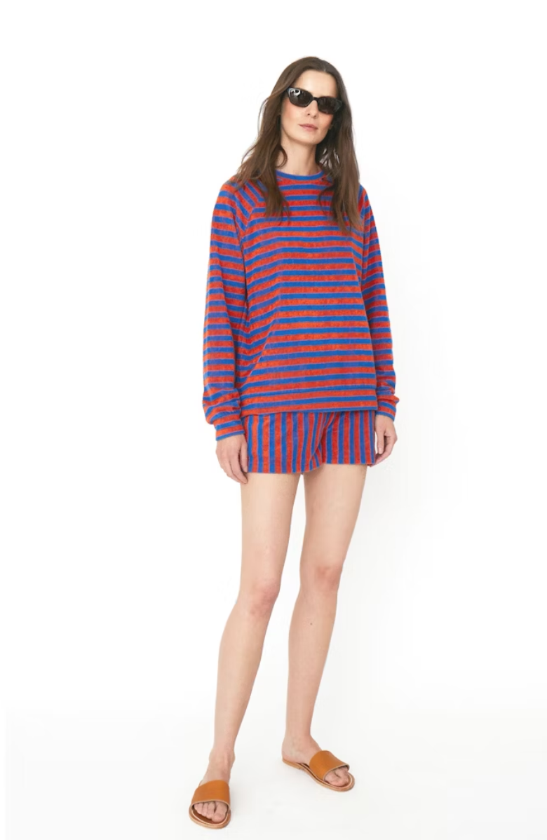 A woman wearing a Kule striped blue and red cotton-blend Terry Franny sweatshirt and shorts combo, accessorized with sunglasses and brown sandals, standing against a white background.
