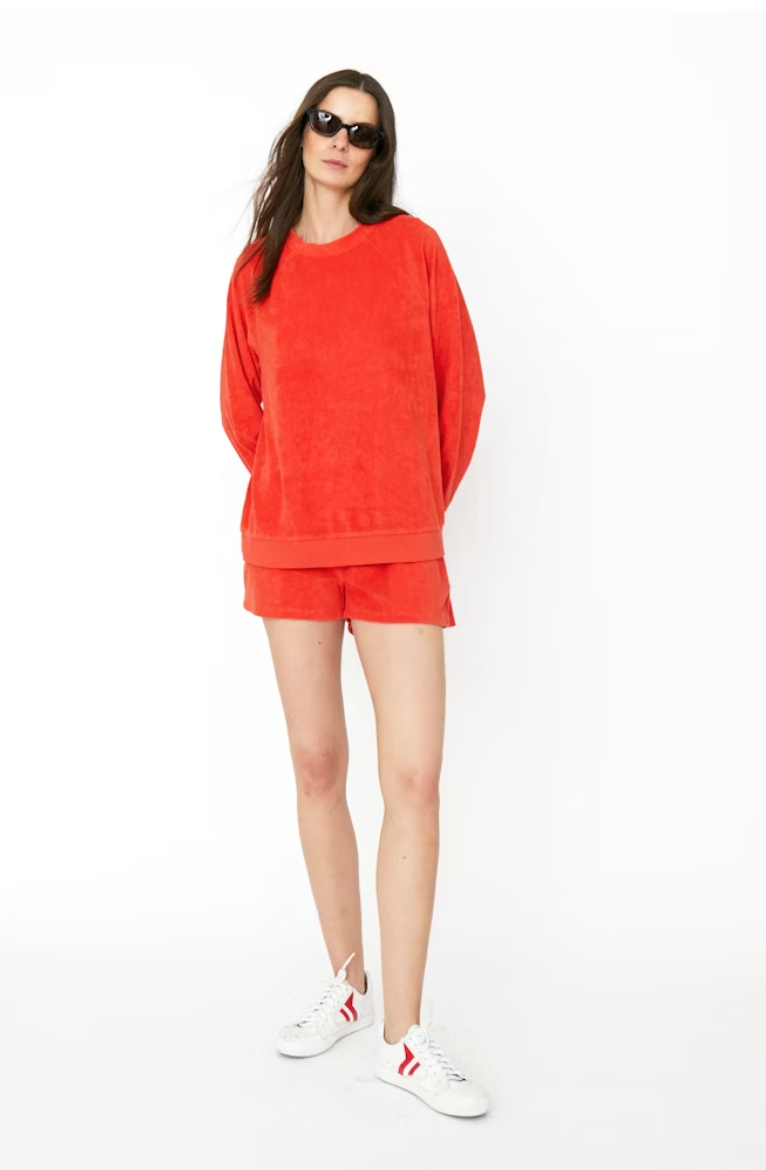 A woman wearing a vibrant red oversized, cotton-blend Terry Franny sweatshirt with matching shorts stands confidently, accessorized with dark sunglasses and white sneakers with red accents. She poses on a plain white background. (Kule)