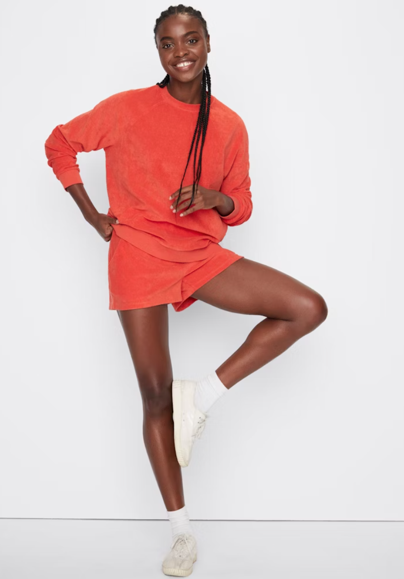 A Black woman with braided hair wearing a Kule Terry Franny sweatshirt and shorts set in orange, along with white sneakers, playfully poses with one leg lifted against a white background.