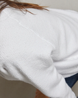 Close-up view of a person wearing a Perfectwhitetee Ziggy Reverse Fleece LS Shrunken Crewneck Sweatshirt and dark jeans, seated outside a bungalow with their back turned partially to the camera. The focus is on the texture of the sweatshirt.