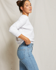 A woman in a white Ziggy Reverse Fleece LS Shrunken Crewneck Sweatshirt and blue jeans looks over her shoulder with a slight smile against the neutral background of a bungalow. She has her hair in a bun and adopts a casual pose.