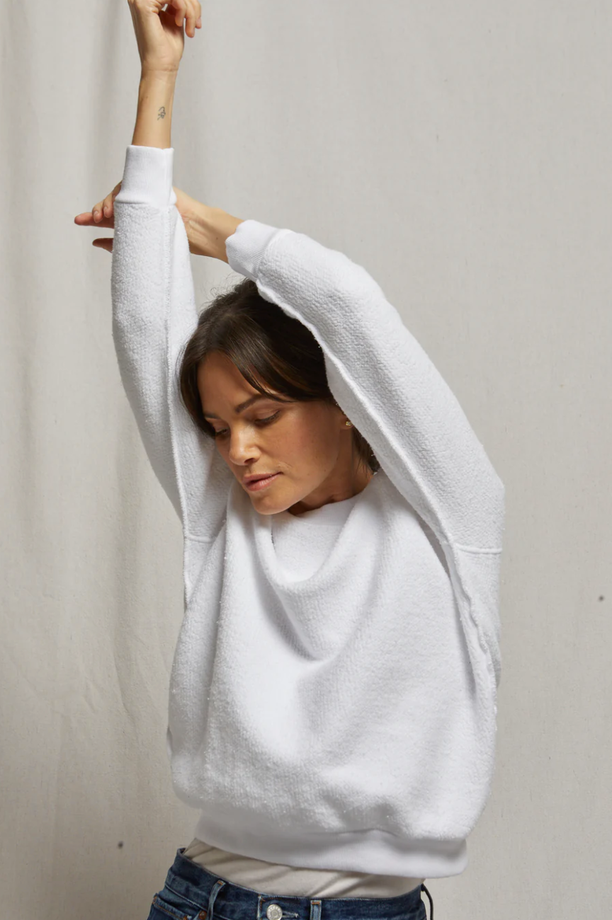 A woman in a white Ziggy Reverse Fleece LS Shrunken Crewneck Sweatshirt and jeans stretches one arm upwards, looking down and standing against a plain beige background in a bungalow.