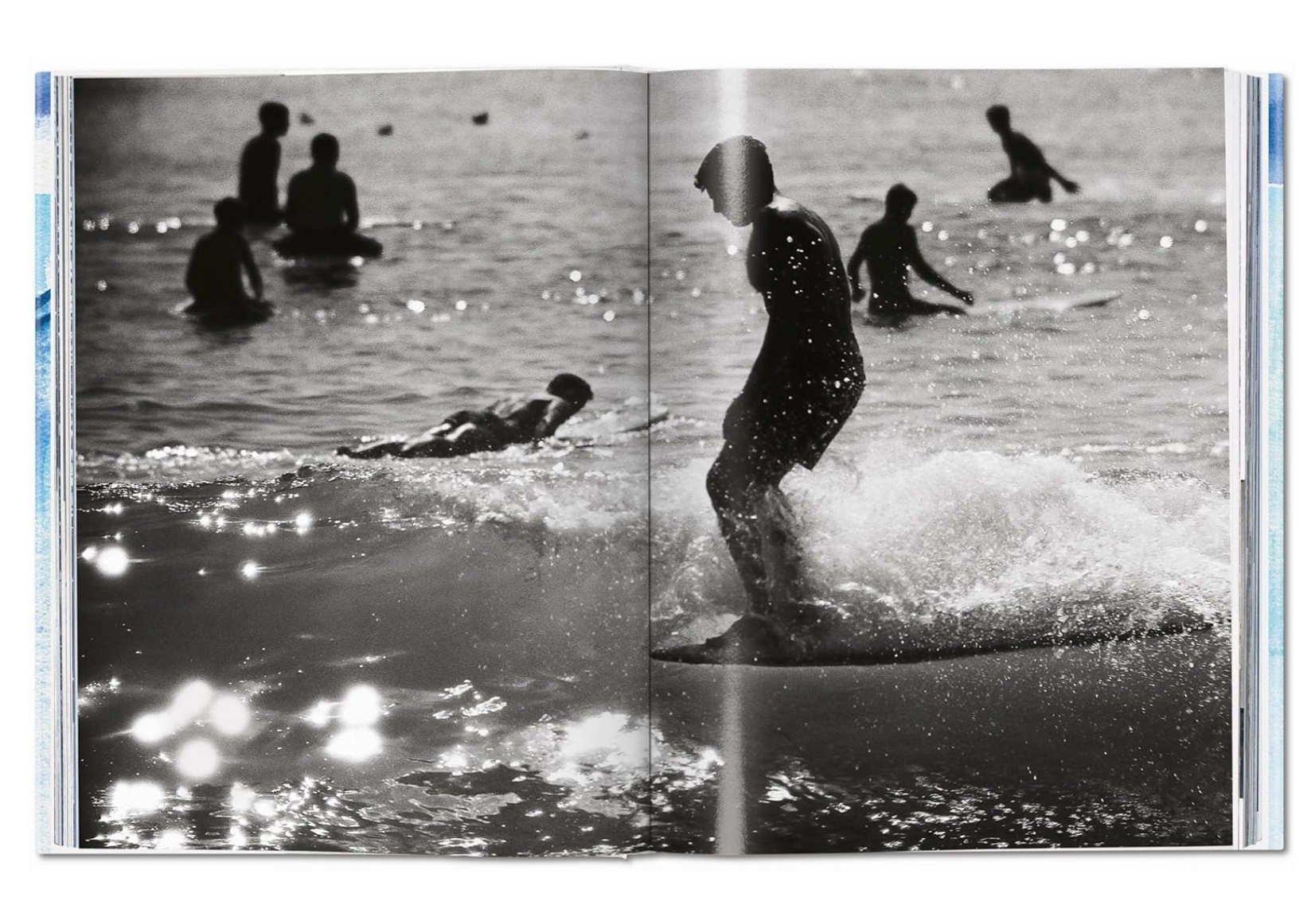 A black and white photo showing multiple people enjoying a day at the beach, with one person in focus seen running through shallow water near a bungalow, sunlight reflecting off the surface from the Breitling Book of Surfing by Random House.