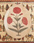 An oval-shaped ceramic plate decorated with a design of three red poppies against a Faire-style tablecloth featuring ancient figures and hieroglyphics.