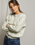 A woman with long brown hair stands against a neutral background in her bungalow, wearing a cozy Ziggy Reverse Fleece LS Shrunken Crewneck Sweatshirt from Perfectwhitetee and blue jeans, arms crossed and looking at the camera.