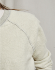 Close-up of a person wearing a Perfectwhitetee Ziggy Reverse Fleece LS Shrunken Crewneck Sweatshirt with visible texture and seams, emphasizing details in the fabric. The background is plain and neutral-colored, echoing the simplicity of a bungalow.