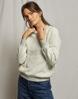 A woman in a Perfectwhitetee Ziggy Reverse Fleece LS Shrunken Crewneck Sweatshirt and jeans poses against a neutral background in a bungalow, lightly touching her collar with a subtle smile.