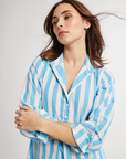 A woman with shoulder-length brown hair wearing a blue and white striped button-up shirt looks thoughtfully to the side on the porch of her Scottsdale, Arizona bungalow, her hair blowing slightly in the breeze wearing a Sadie Long Caftan by Mille.