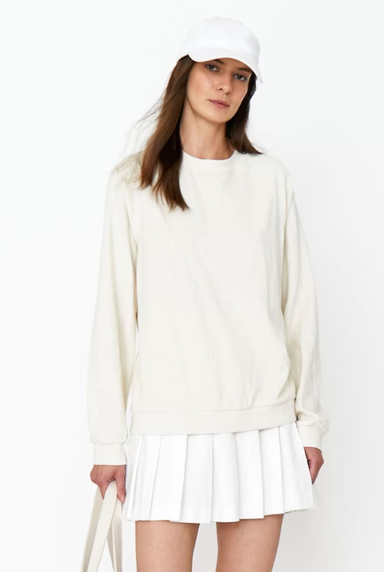 A woman in a stylish white outfit including a cap, Kule's Terry Franny sweatshirt, and pleated skirt, paired with a shoulder bag, posing against a plain background.
