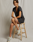 A woman with shoulder-length brown hair, wearing a Perfectwhitetee Opal Japanese Jersey dress and silver sneakers, smiles while sitting on a wooden stool against a beige backdrop in a cozy bungalow.