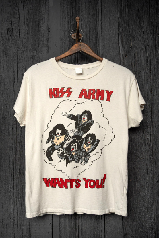 A cream-colored T-shirt with &quot;KISS ARMY WANTS YOU!&quot; printed on it, featuring an illustration of the band members from KISS in a cloud-like outline, hanging on a rustic wooden wall in a bungalow-style room by Made Worn.