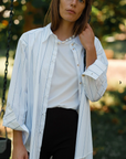 A woman in a white The Stripe Pop Shirt by Donni and a white T-shirt stands near a swing, touching her hair and looking intently at the camera. She wears a chunky pearl necklace.