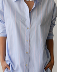 A woman wearing a light blue striped Donni Stripe Pop Shirt with hands on hips. She has a chunky white necklace and the shirt features vertical stripes and wooden buttons.