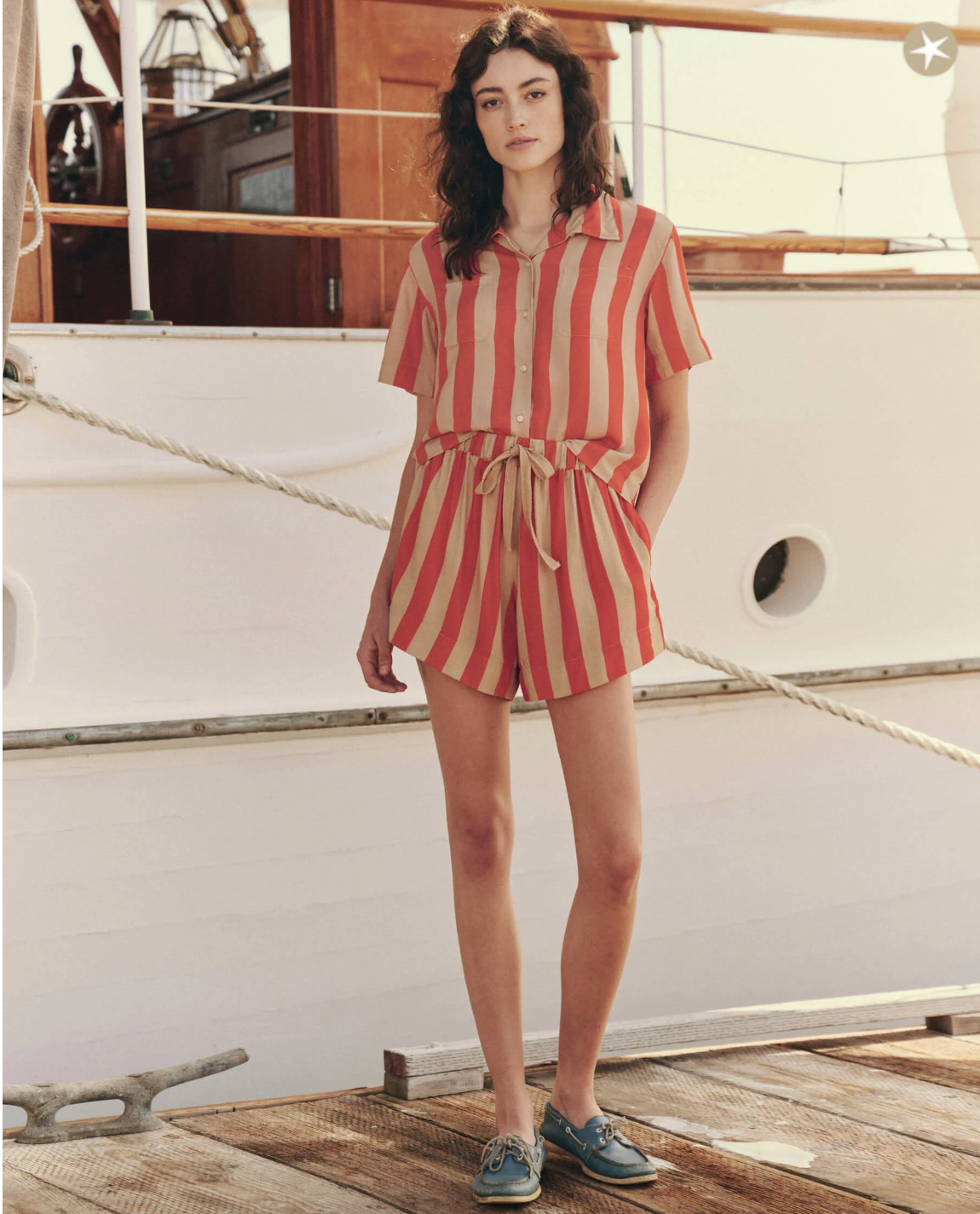 Person with long wavy hair stands on a dock in front of a boat. They wear a matching set with bold, red and beige vertical stripes, consisting of The Bonfire Short from The Great Inc., a short-sleeve button-up shirt, and shorts tied at the adjustable waist. They also wear boat shoes.