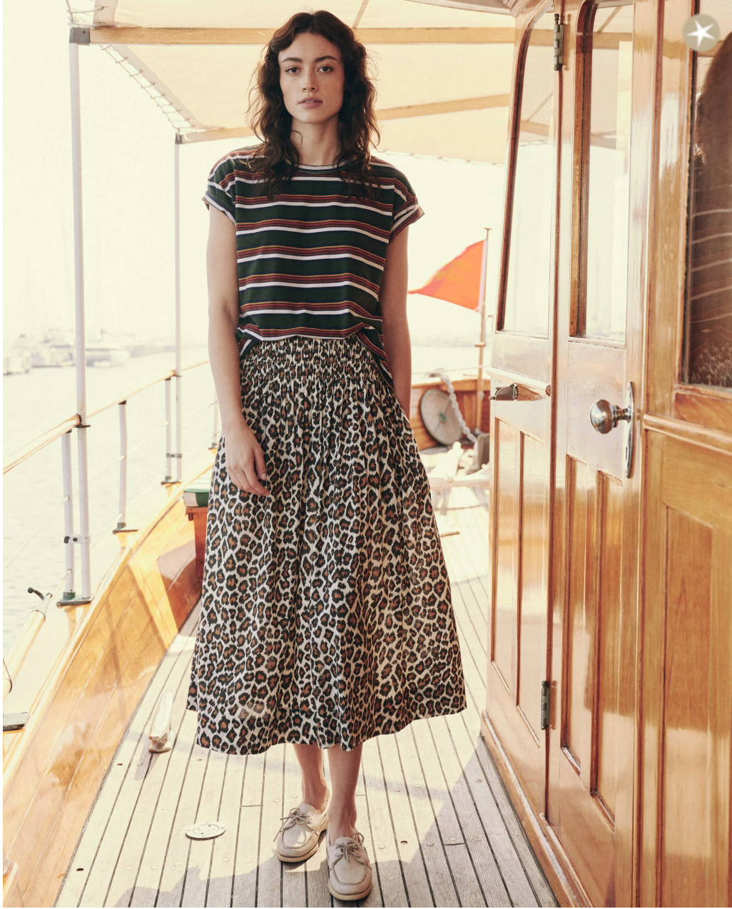 A woman with wavy hair stands on a wooden boat deck. She wears a shadow stripe textured cotton T-shirt, The Viola Skirt by The Great Inc. with a wide smocked waistband, and white sneakers. The boat has a polished wooden cabin door and a shaded canopy area. The background is bright and sunlit.
