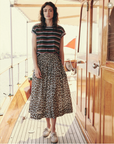 A woman with wavy hair stands on a wooden boat deck. She wears a shadow stripe textured cotton T-shirt, The Viola Skirt by The Great Inc. with a wide smocked waistband, and white sneakers. The boat has a polished wooden cabin door and a shaded canopy area. The background is bright and sunlit.
