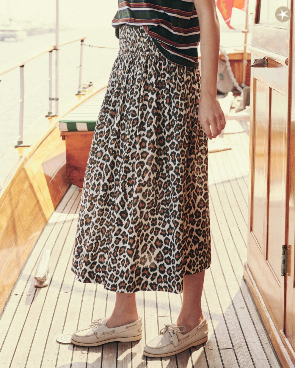 A person stands on a wooden deck wearing a leopard print skirt, a striped shirt, and light-colored boat shoes. The person's face is not visible. In the background, there are wooden elements of what appears to be a boat. The outfit features The Viola Skirt by The Great Inc. with its wide smocked waistband.