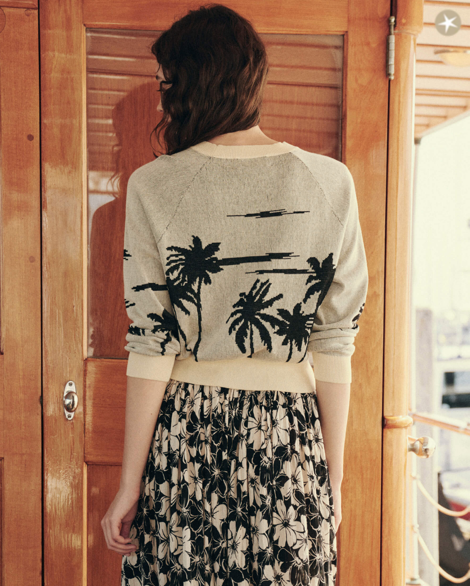 A person with wavy brown hair stands with their back to the camera, wearing The Palm Pullover by The Great Inc. in beige paired with a black and white floral skirt. Wooden doors with brass fixtures are in the background, illuminated by some light that seeps through.