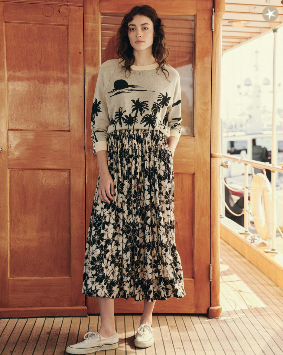 A woman with long, wavy hair stands on a wooden deck in front of a door. She is wearing **The Palm Pullover by The Great Inc.**, paired with a black and white floral skirt and beige boat shoes. The background includes part of a boat's exterior.