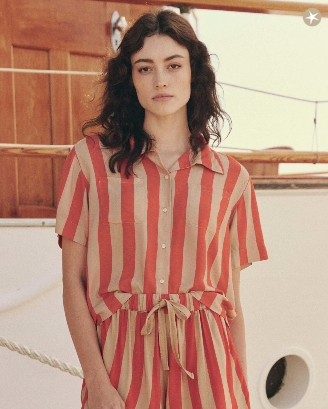 A person with long, wavy hair is wearing The Bowling Shirt by The Great Inc., a vintage men's shirt with a short-sleeve, button-down style and matching pants featuring wide red and beige vertical stripes. They are standing in front of a wooden structure and rope on a boat.
