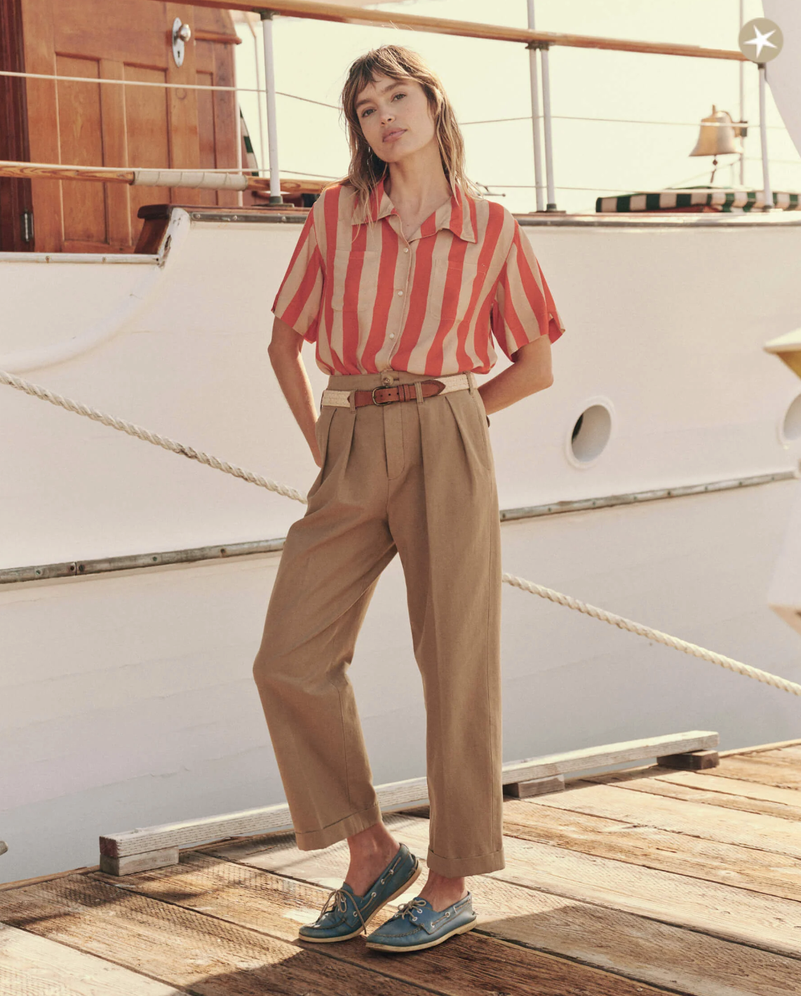 A woman stands on a wooden deck in front of a boat, wearing a short-sleeved, red and white striped The Bowling Shirt by The Great Inc. tucked into high-waisted, tan trousers. Her relaxed fit outfit is completed with teal boat shoes. She gazes off to the side with a relaxed expression.