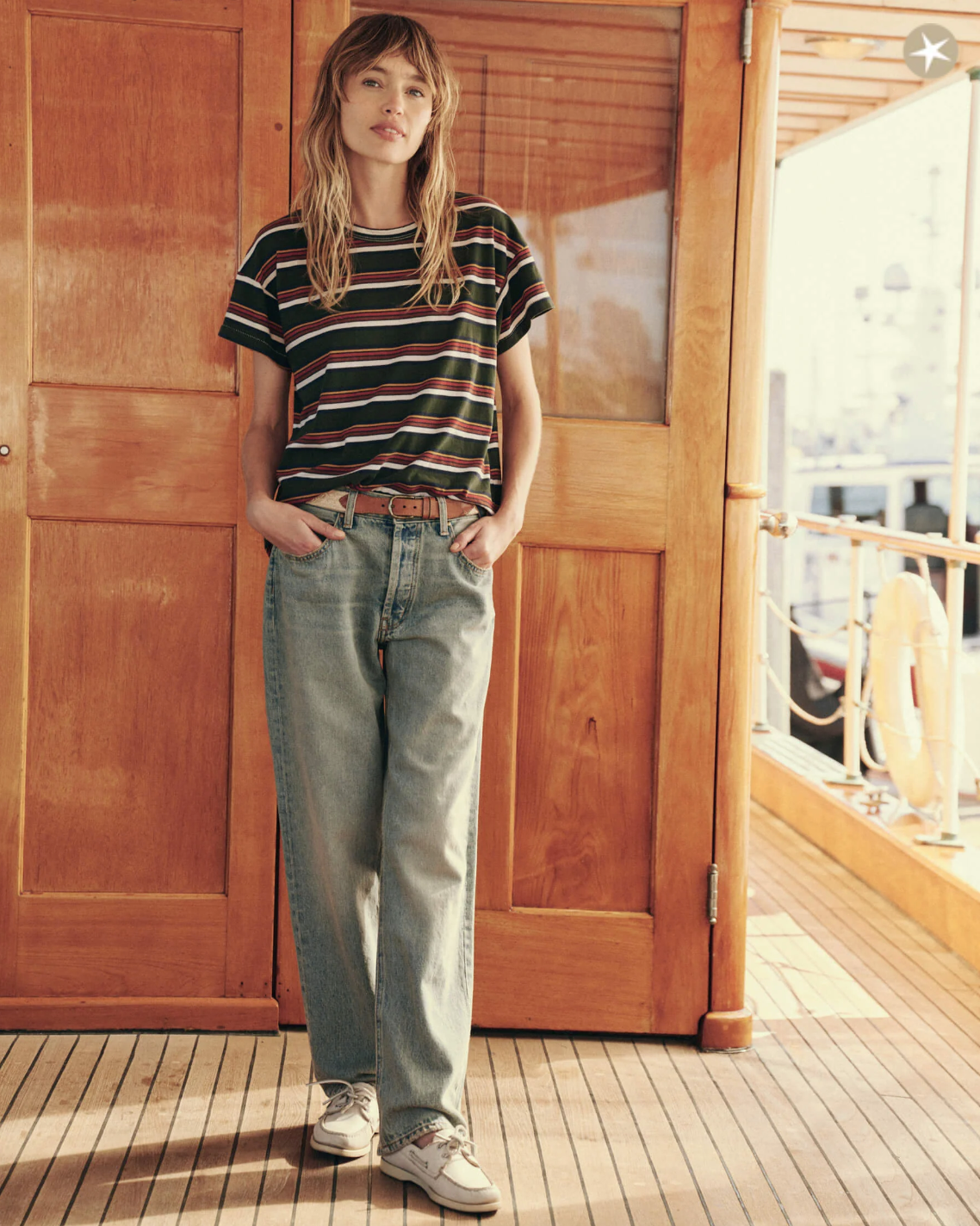 A person with long hair stands on a wooden deck in front of a wooden door. They are wearing The Boxy Crew by The Great Inc., high-waisted jeans, and sneakers, with hands in pockets. Sunlight filters through, casting a warm glow on the scene. The vintage tee adds to the charm of this handcrafted locally scene.