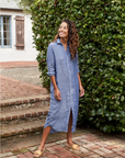 A person smiles while standing outside on a brick pathway, dressed in a long, light blue Frank & Eileen Rory Shirtdress S24. The loose fit of the 100% linen fabric flows gracefully to maxi-length, complemented by beige sandals. Green foliage and part of a white house with brown shutters are visible in the background.