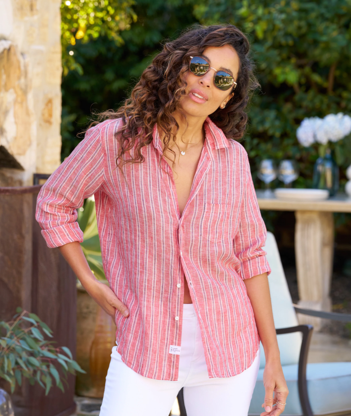 A person with long, curly hair is standing outdoors, wearing sunglasses and a relaxed fit Eileen Shirt by Frank & Eileen paired with white pants. They are slightly smiling, with one hand in their pocket. There are plants and outdoor furniture in the background.