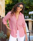 A person with long, curly hair is standing outdoors, wearing sunglasses and a relaxed fit Eileen Shirt by Frank & Eileen paired with white pants. They are slightly smiling, with one hand in their pocket. There are plants and outdoor furniture in the background.