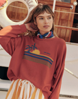 A woman stands on a boat deck, wearing The Teammate Sweatshirt by The Great Inc., with a sunset and palm tree design, and a striped scarf around her neck. She has blonde hair with bangs and is resting one hand on the back of her head. The handcrafted sweatshirt, made in the U.S.A., perfectly complements the background with its life ring.