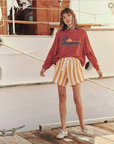 A woman stands on a wooden dock next to a boat. She is wearing The Teammate Sweatshirt by The Great Inc. with a palm tree design, yellow and white striped shorts with a drawstring, and white shoes. She has a blue scarf around her neck and her hands hang loosely by her sides.