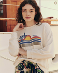 A person with wavy brown hair is standing on a boat, resting their left hand on their neck. They're wearing The Teammate Sweatshirt by The Great Inc., which is cream-colored and has a vintage sunset and palm tree design, made in the U.S.A., and multicolored striped shorts. The background shows boat railings and wooden elements.