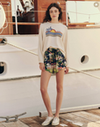 A person with long, wavy hair stands on a wooden deck in front of a boat. They wear The Teammate Sweatshirt by The Great Inc., paired with floral shorts and white sneakers. The background features part of the boat's cabin and a railing.