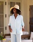 A woman wearing a straw hat and a light blue striped Frank & Eileen Eileen Shirt stands outdoors. She is smiling with her hands relaxed by her side. In the background, an outdoor seating area with light-colored furniture and a small table adorned with flowers and lemons creates a relaxed fit vibe.