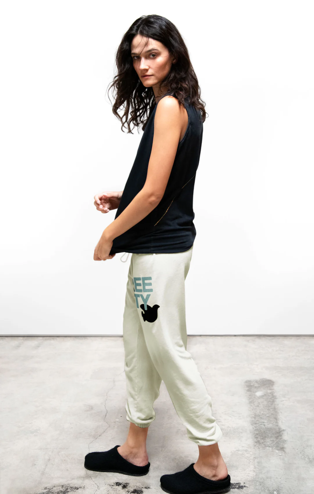 A woman stands on a concrete floor in front of a plain white wall. She has long curly hair and is wearing a sleeveless black top and FREECITY Large Sweatpant by Free City (sparrow, LLC) with a graphic of a bird and the word "TOGETHER" partially visible near the pocket. She is wearing black slippers.