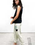 A woman stands on a concrete floor in front of a plain white wall. She has long curly hair and is wearing a sleeveless black top and FREECITY Large Sweatpant by Free City (sparrow, LLC) with a graphic of a bird and the word "TOGETHER" partially visible near the pocket. She is wearing black slippers.