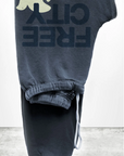 A pair of dark blue, unisex Free City (sparrow, LLC) FREECITY Large Sweatpant is hung, showcasing the leg section prominently. Made from lightweight French terry, these sweatpants feature an inverted FREECITY print in large bold letters along with a graphic of a yellow bird. There is a drawstring at the waistband.