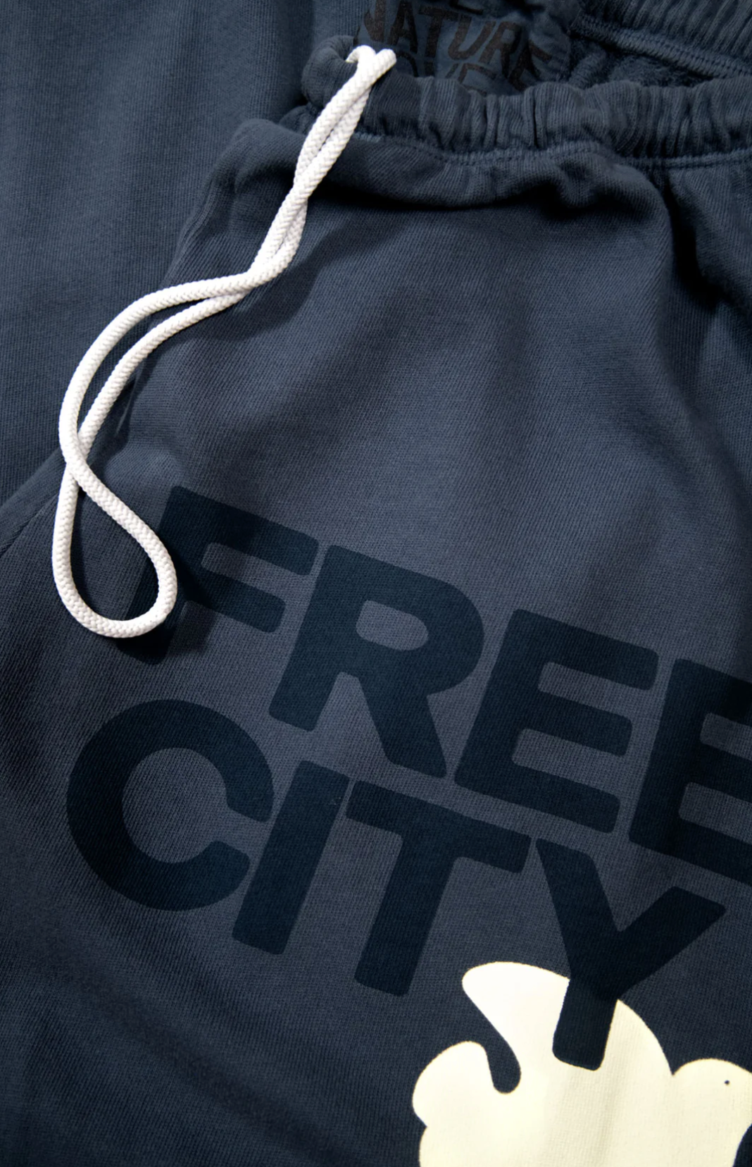 Close-up image of a pair of dark gray Free City (sparrow, LLC) FREECITY Large Sweatpant featuring a white drawstring and the text &quot;FREE CITY&quot; in bold black letters. A partially visible white graphic is positioned below the text, all crafted from lightweight French terry fabric.