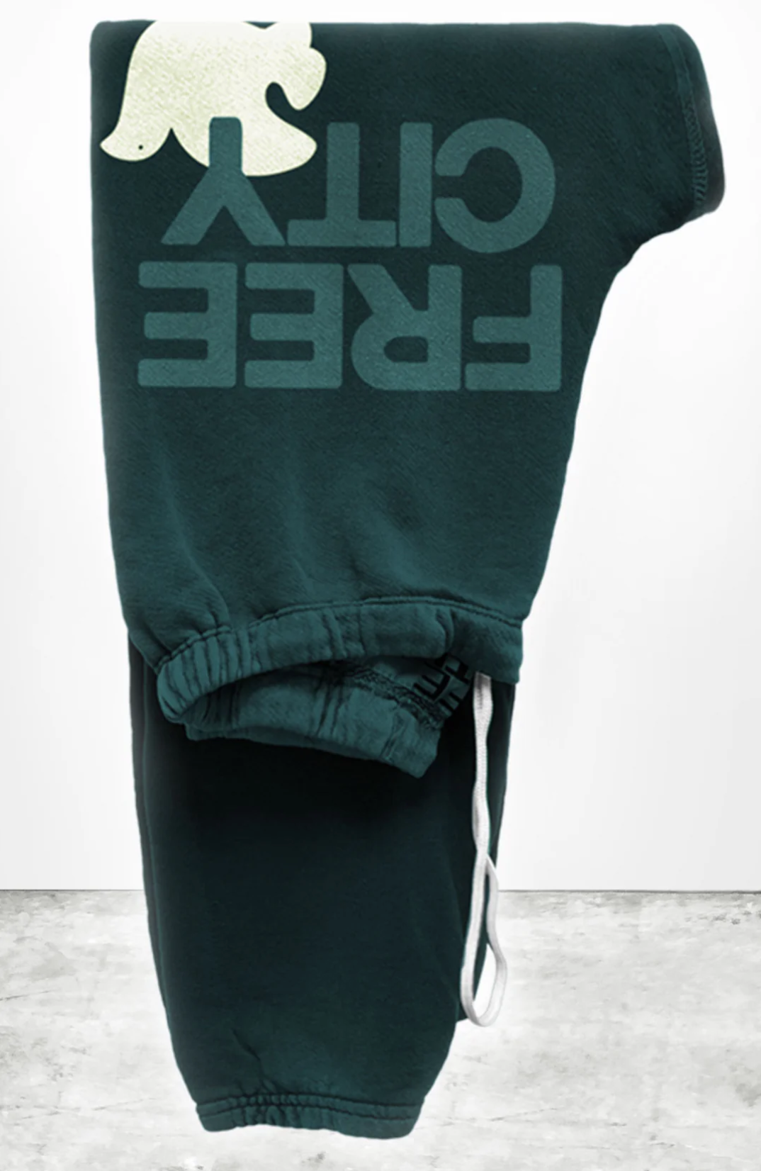 A pair of dark green sweatpants folded neatly. These unisex Free City (sparrow, LLC) FREECITY Large Sweatpant feature a white bear graphic and bold, upside-down "FREE CITY" print in dark teal. Made from lightweight French terry, the drawstring waist is slightly visible with a white drawstring hanging loosely.