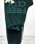 A pair of dark green sweatpants folded neatly. These unisex Free City (sparrow, LLC) FREECITY Large Sweatpant feature a white bear graphic and bold, upside-down "FREE CITY" print in dark teal. Made from lightweight French terry, the drawstring waist is slightly visible with a white drawstring hanging loosely.