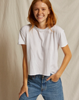 A person with long, red hair smiles while wearing a Perfectwhitetee Harley SS Boxy Crew and high-waisted blue jeans. They have one hand in their pocket and stand against a simple, light-colored background, exuding a relaxed bungalow style.
