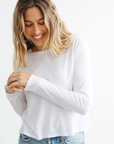 A person with wavy, shoulder-length blond hair is smiling and looking down while wearing a Perfectwhitetee kitt knit in white paired with light blue jeans. The background is plain white.