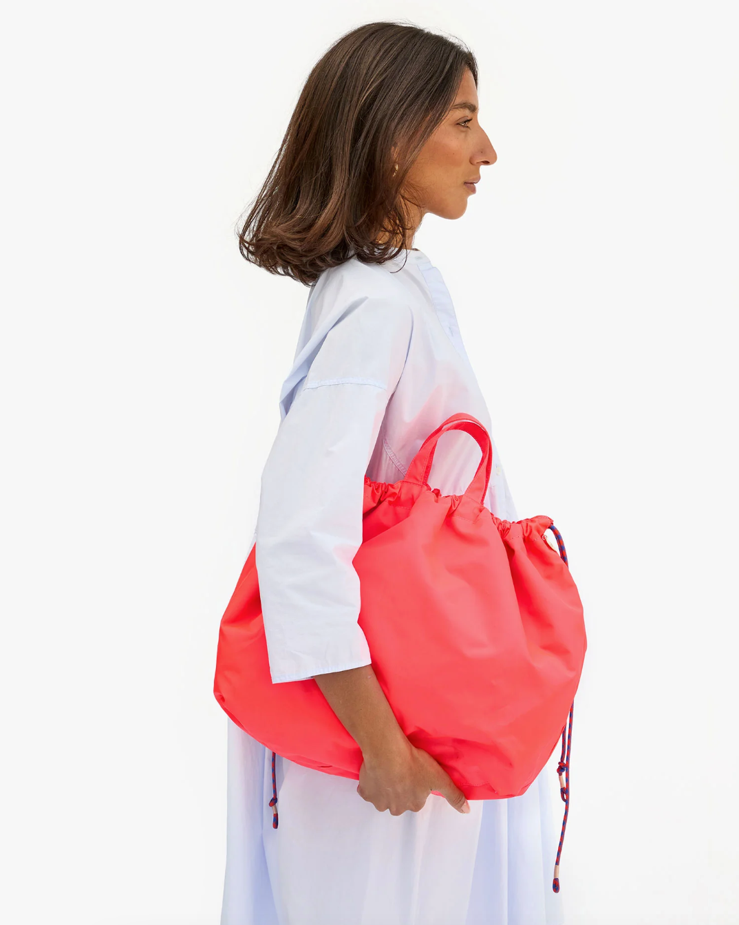 A woman with shoulder-length dark hair is standing sideways, showcasing her style with a bright red Sac Tout from Clare Vivier. She is wearing a light blue long-sleeved dress. The background is white, emphasizing the simplicity and elegance of her look.