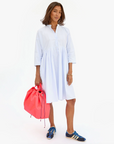 A person stands against a white background wearing a stylish white knee-length dress with a collar and three-quarter sleeves. They carry a large, bright pink Sac Tout by Clare Vivier and wear blue sneakers with white stripes. With shoulder-length brown hair, they accessorize with jewelry that has an Arizona flair.