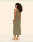 A woman with curly hair is standing and facing away. She is wearing an olive-green Pia Xirena Dress by Xirena that reaches mid-calf and beige sandals. The minimalistic scene, reminiscent of an Arizona bungalow style, features a plain cream background.